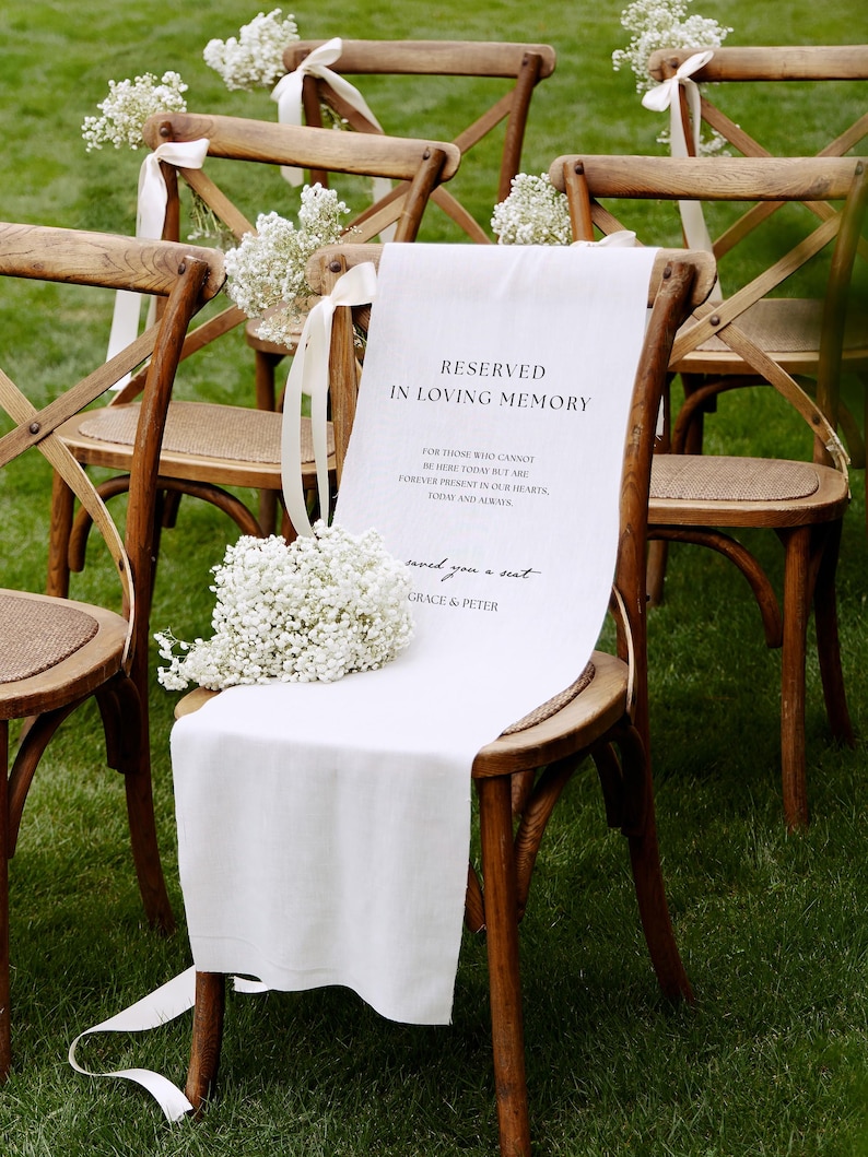 May include: A wooden cross-back chair draped with a white fabric sign reading RESERVED IN LOVING MEMORY, FOR THOSE WHO CANNOT BE HERE TODAY BUT ARE FOREVER PRESENT IN OUR HEARTS, TODAY AND ALWAYS, saved you a seat, GRACE & PETER, with baby's breath.
