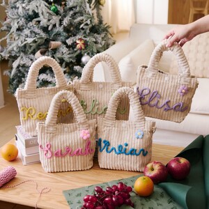 May include: Five beige corduroy tote bags with embroidered names in various colors. Each bag has a matching corduroy handle and a small decorative flower. The bags are displayed on a wooden table with fruit and wrapping supplies.