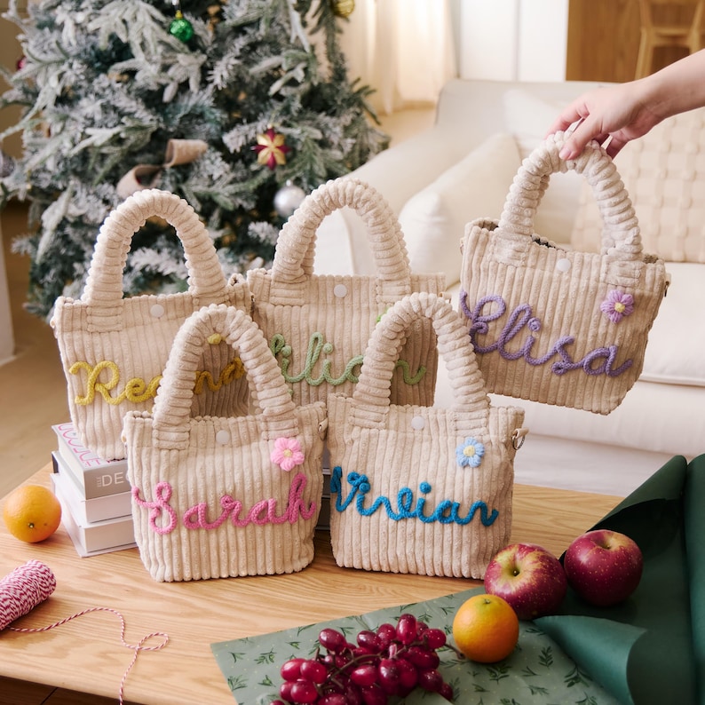 May include: Five beige corduroy tote bags with embroidered names in various colors. Each bag has a matching corduroy handle and a small decorative flower. The bags are displayed on a wooden table with fruit and wrapping supplies.