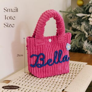 May include: A pink corduroy tote bag with the name "Bella" in blue script. The bag has a rounded shape, a button closure, and a small blue flower. It measures 21cm high and 22cm wide.