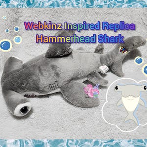 Op de afbeelding: Een grijs pluche hamerhaai-speeltje met een brede kop, donkere ogen en zijvinnen. De tekst Webkinz Inspired Replica Hammerhead Shark is toegevoegd. Een kleine cartoon-haai-illustratie en blauwe bubbelafbeeldingen zijn op de ondergrond zichtbaar.