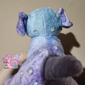 Puede incluir: Un peluche en tonos azules y morados, con cola y orejas. El juguete est&aacute; decorado con adornos transparentes y brillantes. La parte trasera del juguete es visible y parece estar sostenido por una persona.