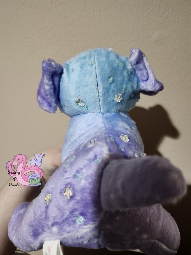 Puede incluir: Un peluche en tonos azules y morados, con cola y orejas. El juguete est&aacute; decorado con adornos transparentes y brillantes. La parte trasera del juguete es visible y parece estar sostenido por una persona.