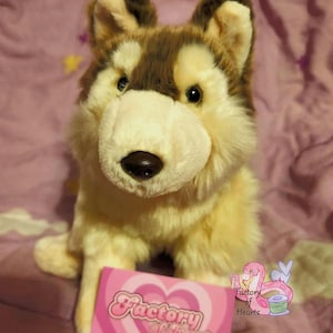 May include: A plush dog toy with brown and cream-colored fur, pointed ears, and a dark nose. A rectangular pink card with a heart pattern and the text "Factory of Hearts" is positioned in front of the plush, along with a small logo of a pink snake.