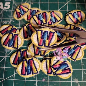 Può includere: Una collezione di toppe ricamate con bordo giallo e sfondo bianco. Ogni toppa presenta un colorato design "W" in rosso, blu e giallo. Le toppe sono su un tappetino da taglio verde.