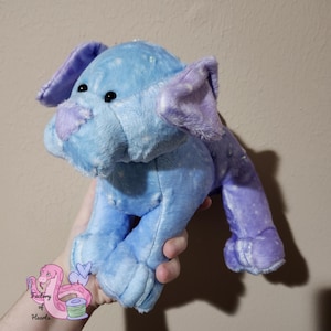 Puede incluir: Un elefante de peluche en tonos azules y morados. El elefante tiene orejas grandes, una trompa y ojos negros peque&ntilde;os. El peluche es suave y tierno, con una superficie texturizada. El elefante est&aacute; siendo sostenido por una persona.