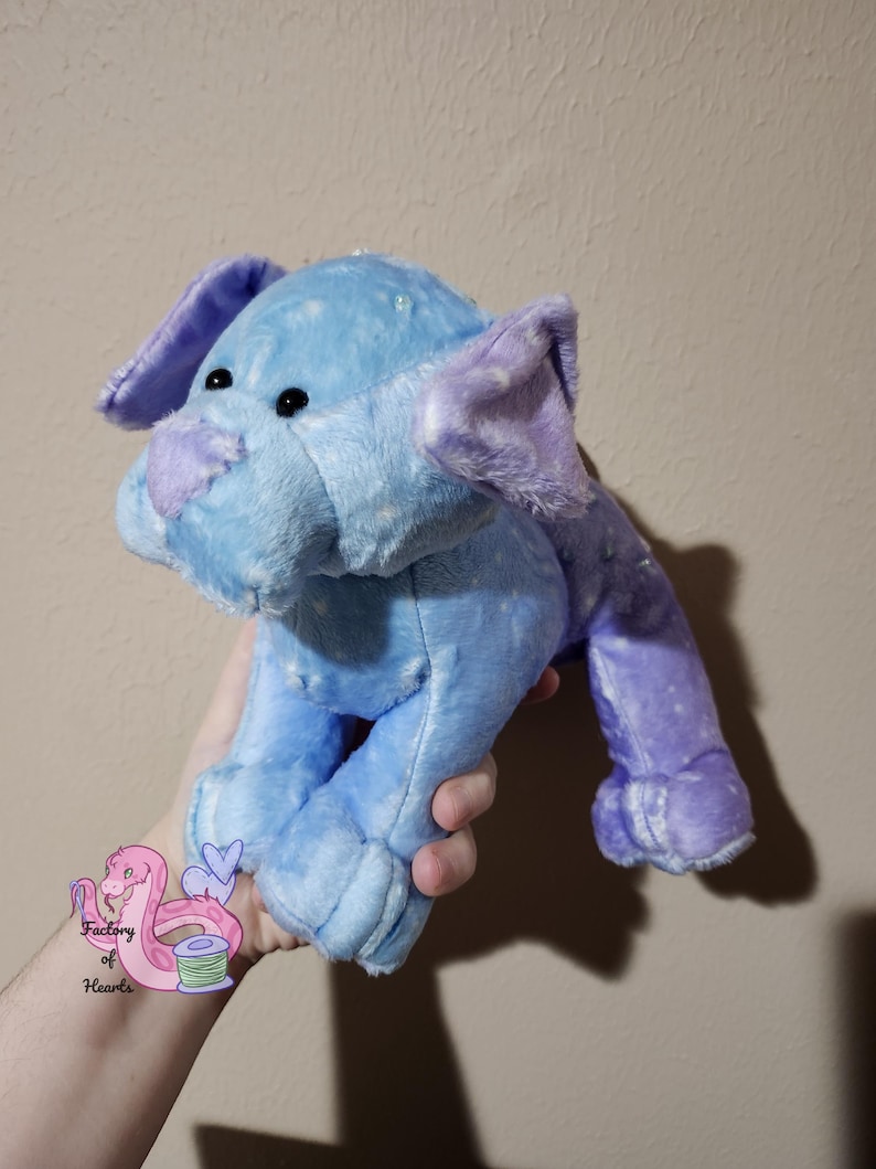 Puede incluir: Un elefante de peluche en tonos azules y morados. El elefante tiene orejas grandes, una trompa y ojos negros peque&ntilde;os. El peluche es suave y tierno, con una superficie texturizada. El elefante est&aacute; siendo sostenido por una persona.