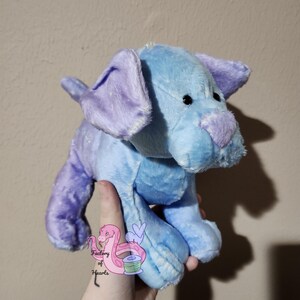 Puede incluir: Un peluche con forma de perro en tonos azules y morados. El perro tiene orejas moradas ca&iacute;das, una nariz morada y un cuerpo azul claro. El juguete se sostiene en una mano, con un peque&ntilde;o logotipo visible en la mano.