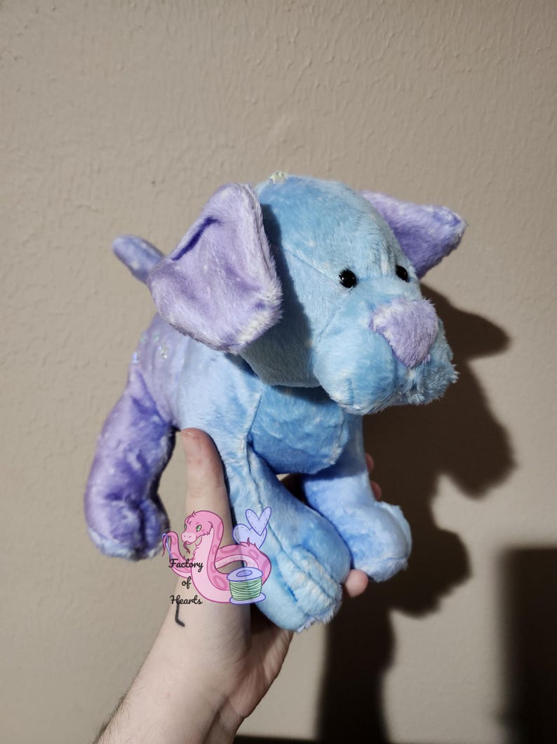Puede incluir: Un peluche con forma de perro en tonos azules y morados. El perro tiene orejas moradas ca&iacute;das, una nariz morada y un cuerpo azul claro. El juguete se sostiene en una mano, con un peque&ntilde;o logotipo visible en la mano.