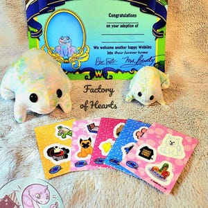 May include: Certificate of Adoption for a Webkinz plush toy. The certificate is green and yellow with a picture of a frog. The plush toy is a light blue and white frog. There are six small stickers with different Webkinz characters on them.