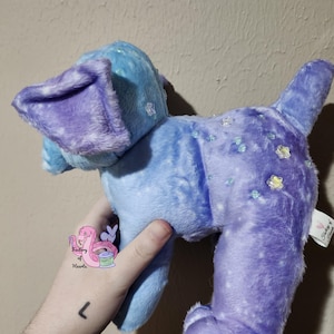 Puede incluir: Un perro de peluche en tonos azules y morados. El perro tiene orejas grandes y ca&iacute;das y una cola. El peluche est&aacute; decorado con peque&ntilde;os adornos brillantes. El juguete es suave y esponjoso, con una etiqueta.