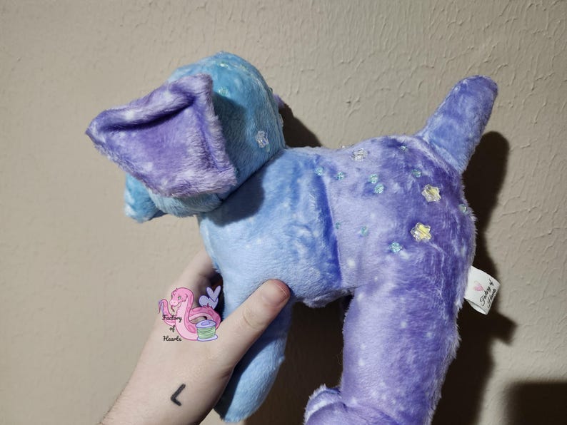 Puede incluir: Un perro de peluche en tonos azules y morados. El perro tiene orejas grandes y ca&iacute;das y una cola. El peluche est&aacute; decorado con peque&ntilde;os adornos brillantes. El juguete es suave y esponjoso, con una etiqueta.