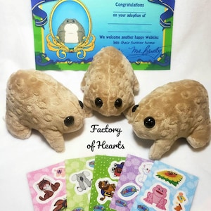 May include: Three brown plush Webkinz frogs with textured fur. The frogs are sitting on a white surface. There is a certificate of adoption for a Webkinz frog, a sticker sheet with a koala, a lion, a cat, and a frog, and a sign that says "Factory of Hearts".