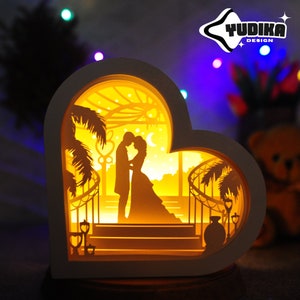 May include: A white heart-shaped paper lamp with a 3D design of a couple standing on a staircase in a garden setting. The lamp is illuminated from within, casting a warm glow on the design. The design includes palm trees, a moon, and stars.