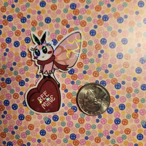 Peut inclure: Un autocollant holographique rose et blanc représentant un papillon de nuit tenant une boîte en forme de cœur rouge avec le texte "Bee Mine".