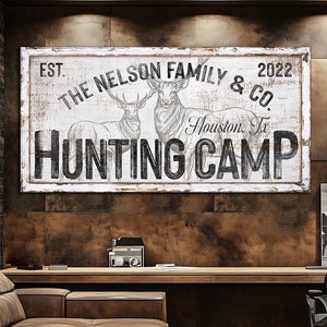 May include: A white sign with black text and a drawing of two deer. The sign reads "EST. THE NELSON FAMILY & CO. 2022 Houston, Tx HUNTING CAMP".