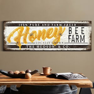 Op de afbeelding: Een rustiek wit bord met zwarte letters met de tekst "100% Zuiver en vers van de boerderij Honey Bee Farm - Lokaal geteeld - The Hudson & Co.". Het woord "Honey" staat in een gestileerd lettertype met een druppelend honing-effect en een honingraatpatroon. Er vliegen kleine bijen rond het woord "Honey".