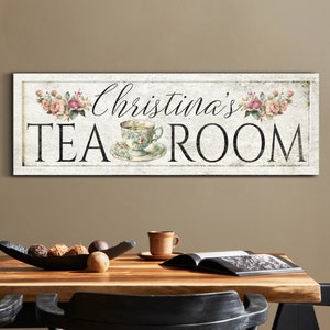 May include: A white sign with a distressed look featuring a teacup and the text "Christina's Tea Room" with pink flowers on either side.