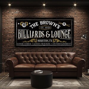 May include: A black and white sign with gold accents that reads "The Brown's Billiards & Lounge" with the text "Est. 2022" and "Houston, TX" below. The sign also has two billiard balls with the numbers 7 and 8 on them. The sign is hanging on a brick wall above a brown leather sofa.