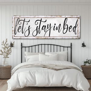 Op de afbeelding: Een wit houten bord met zwarte letters met de tekst "let's stay in bed". Het bord hangt boven een wit bed met een zwart metalen hoofdeinde.