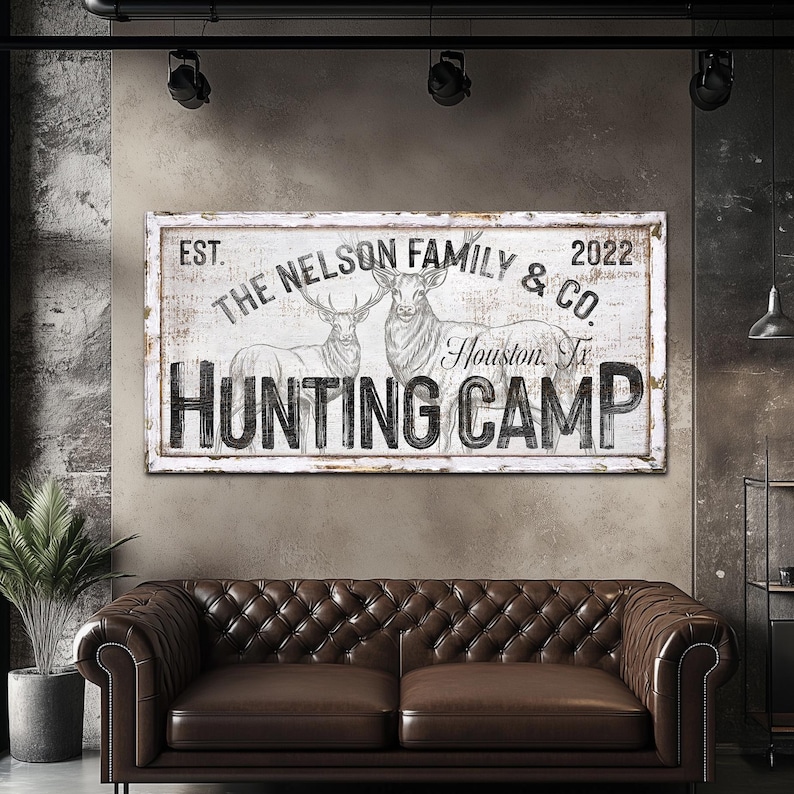 May include: A white wooden sign with black text that reads "EST. THE NELSON FAMILY & CO. 2022 Houston, Tx HUNTING CAMP". The sign features a black and white illustration of two deer with antlers.
