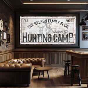 May include: A rustic wooden sign with white paint and black lettering reads "EST. THE NELSON FAMILY & CO. 2022 Houston, Tx HUNTING CAMP". Two stylized deer are depicted in black and white on the sign.