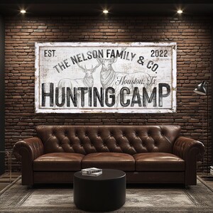 May include: A white sign with black text that reads "EST. THE NELSON FAMILY & CO. 2022 Houston, Tx HUNTING CAMP". The sign features a drawing of two deer in the background.