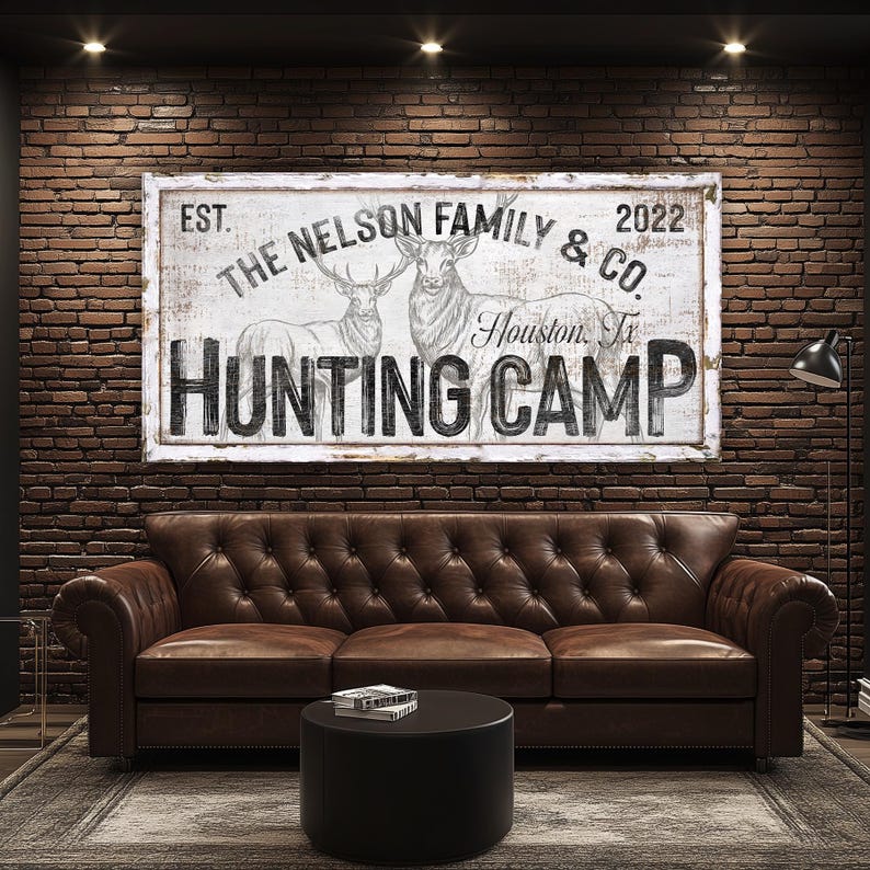May include: A white sign with black text that reads "EST. THE NELSON FAMILY & CO. 2022 Houston, Tx HUNTING CAMP". The sign features a drawing of two deer in the background.