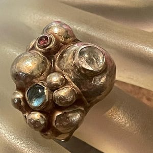 Può includere: Un anello d'argento dal design unico e strutturato, che ricorda delle bolle. L'anello è impreziosito da diverse pietre preziose, tra cui una trasparente, una rossa e una blu. Un gioiello distintivo.
