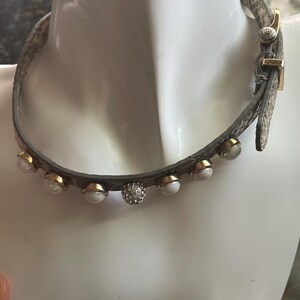 May include: A choker necklace featuring a grey snakeskin-textured band adorned with a series of round white beads set in gold-toned metal, with a single central bead encrusted with clear crystals and a gold-toned buckle closure on the side.