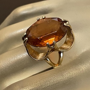May include: A gold-toned ring featuring a large, faceted, amber-colored gemstone. The gemstone is oval-shaped and set in a detailed, openwork gold band. The ring's design includes decorative elements around the gemstone and on the band.