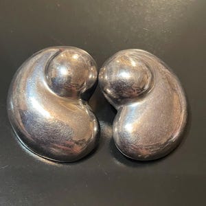 May include: Pair of polished silver-toned earrings. Each earring has a rounded, abstract shape with a smooth, reflective surface. The earrings are displayed on a dark surface, highlighting their metallic sheen.
