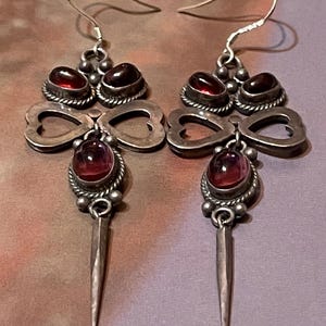 May include: A pair of silver dangle earrings featuring two small oval red stones at the top, a central heart-shaped cutout frame, a larger oval red stone in the middle, and a long, pointed silver spike at the bottom, all suspended from silver hooks.