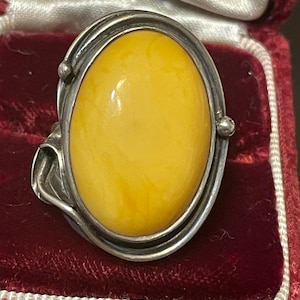 May include: A silver ring with a large, oval amber stone. The amber is a warm yellow colour, set in a detailed silver band. The ring is displayed in a red velvet jewellery box, emphasising its vintage style and design.