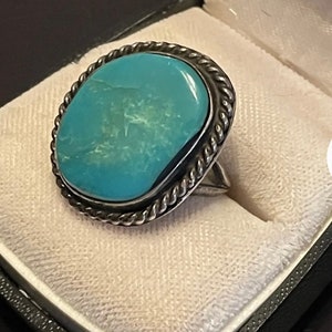 Puede incluir: Un anillo de plata con una gran piedra turquesa engastada en un diseño de cuerda. El anillo está en una caja de anillo blanca.