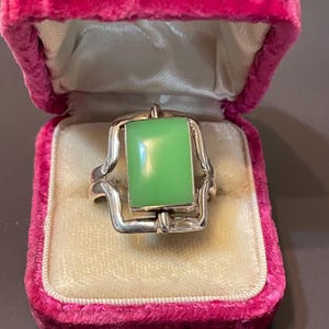 May include: A silver ring featuring a rectangular, smooth, light green stone set within a polished metal frame. The ring is displayed inside a small, open, bright pink velvet-lined jewelry box with a cream-colored interior cushion.