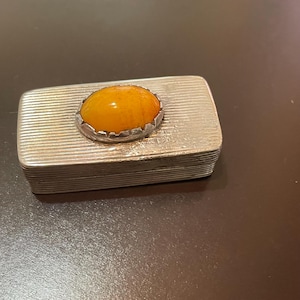 May include: A rectangular silver-toned metal box featuring a textured, horizontally lined surface. Centered on the lid is an oval, smooth, amber-colored stone set within a scalloped, serrated metal bezel that secures the stone to the top.