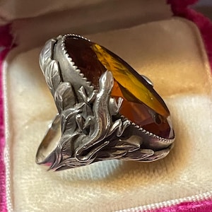 Puede incluir: Un anillo de plata con una gran piedra preciosa de corte ovalado y color ámbar en una banda decorativa. El metal presenta un relieve detallado de un lagarto trepando entre hojas texturizadas que rodean los lados de la piedra central.