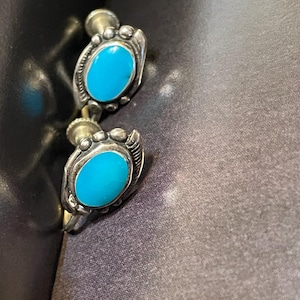Peut inclure: Une paire de boucles d'oreilles à clous argentées avec des pierres ovales turquoise vif. Les boucles d'oreilles présentent un design orné autour des pierres turquoise, avec de petits accents perlés. Les boucles d'oreilles sont posées sur un fond sombre.