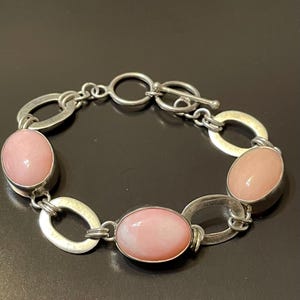 Puede incluir: Una pulsera de plata con piedras preciosas ovaladas de color rosa pálido engastadas en un diseño de eslabones. La pulsera tiene un cierre de palanca y se muestra sobre una superficie oscura. Las piedras son de color pastel.