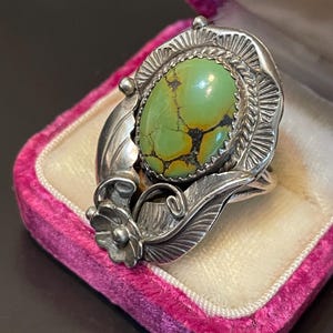 Puede incluir: Un anillo de plata con una piedra ovalada de turquesa verde claro con vetas marrones. La piedra está engastada en un bisel detallado con un diseño de hojas y flores en la banda. El anillo se exhibe en un joyero de terciopelo rosa.