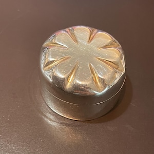Könnte beinhalten: Ein kleiner, runder, silberfarbener Metallbehälter mit Deckel. Die Oberseite des Deckels weist ein erhabenes, geprägtes Blumen- oder Blütenblattmuster auf, das von der Mitte ausstrahlt. Die Seiten des Behälters sind glatt, zylindrisch und reflektierend.