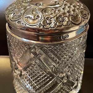 Könnte beinhalten: Ein klares, geschliffenes Glasgefäß mit einem dekorativen, silberfarbenen Deckel. Der Deckel weist florale und Rankenmuster auf. Der Körper des Glases hat ein rautenförmiges Muster. Das zylindrische Glas ist wahrscheinlich ein Behälter.