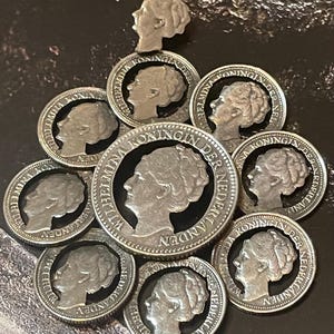 May include: A collection of silver coins and a small silver pin featuring a profile of a woman. The coins are arranged in a circular pattern, with the pin at the top. The coins have text around the edges.