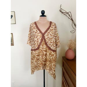 Puede incluir: Una blusa con estampado floral color crema con cuello en V y mangas cortas. La blusa presenta un ribete de encaje marrón a lo largo del escote y el dobladillo. El estampado floral incluye flores marrones y beige.