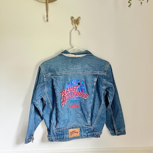 May include: Blue denim jacket with the Planet Hollywood London logo on the back. The logo is a circular design with a pink and blue color scheme. The jacket is hanging on a white hanger.