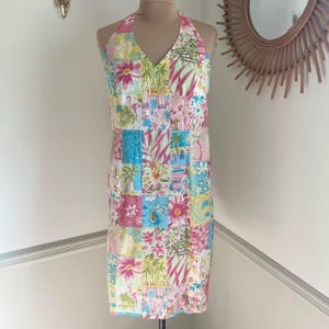 May include: A halter-neck dress with a patchwork design. The dress features a V-neck and a knee-length hemline. The fabric is a mix of floral and tropical patterns in shades of pink, blue, yellow, and green. The dress is displayed on a mannequin.