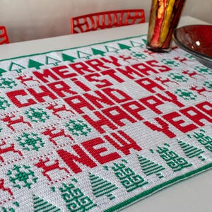 May include: White crochet table runner with a red and green Christmas design. The runner features the words "Merry Christmas and Happy New Year".