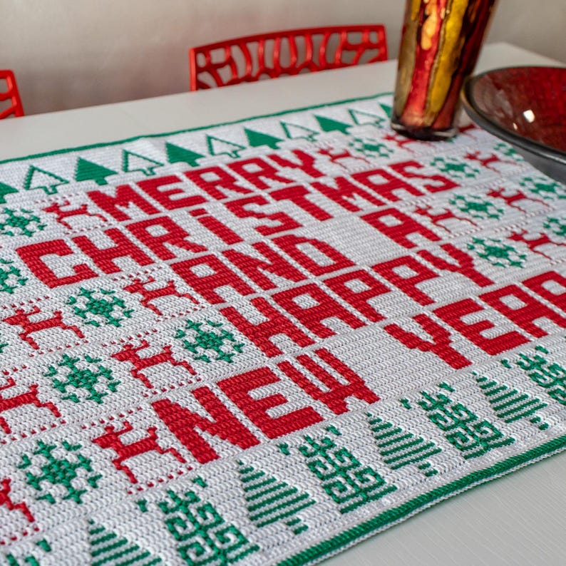 May include: White crochet table runner with a red and green Christmas design. The runner features the words "Merry Christmas and Happy New Year".