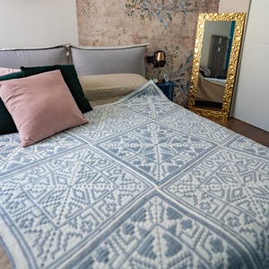 Puede incluir: Una cama cubierta con un edredón azul y blanco estampado. El edredón presenta un diseño geométrico. La cama tiene varias almohadas en tonos rosa, verde y gris. Un espejo con marco dorado se apoya en la pared.